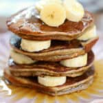 A stack of banana protein pancakes with banana slices and maple syrup.