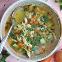 A bowl of gluten free chicken soup with a spoon on a pink background.