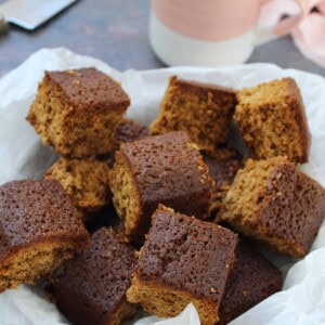 Squares of gluten free parkin in a dish lined with baking paper and a cup in the background.