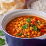 A dish of gluten free chickpea curry with poppadoms and rice behind.