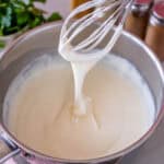 A whisk in a pan of creamy gluten free white sauce, showing how smooth the sauce is with no lumps.