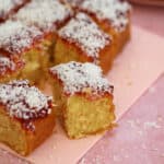 Squares of jam and coconut sponge on a pink board.