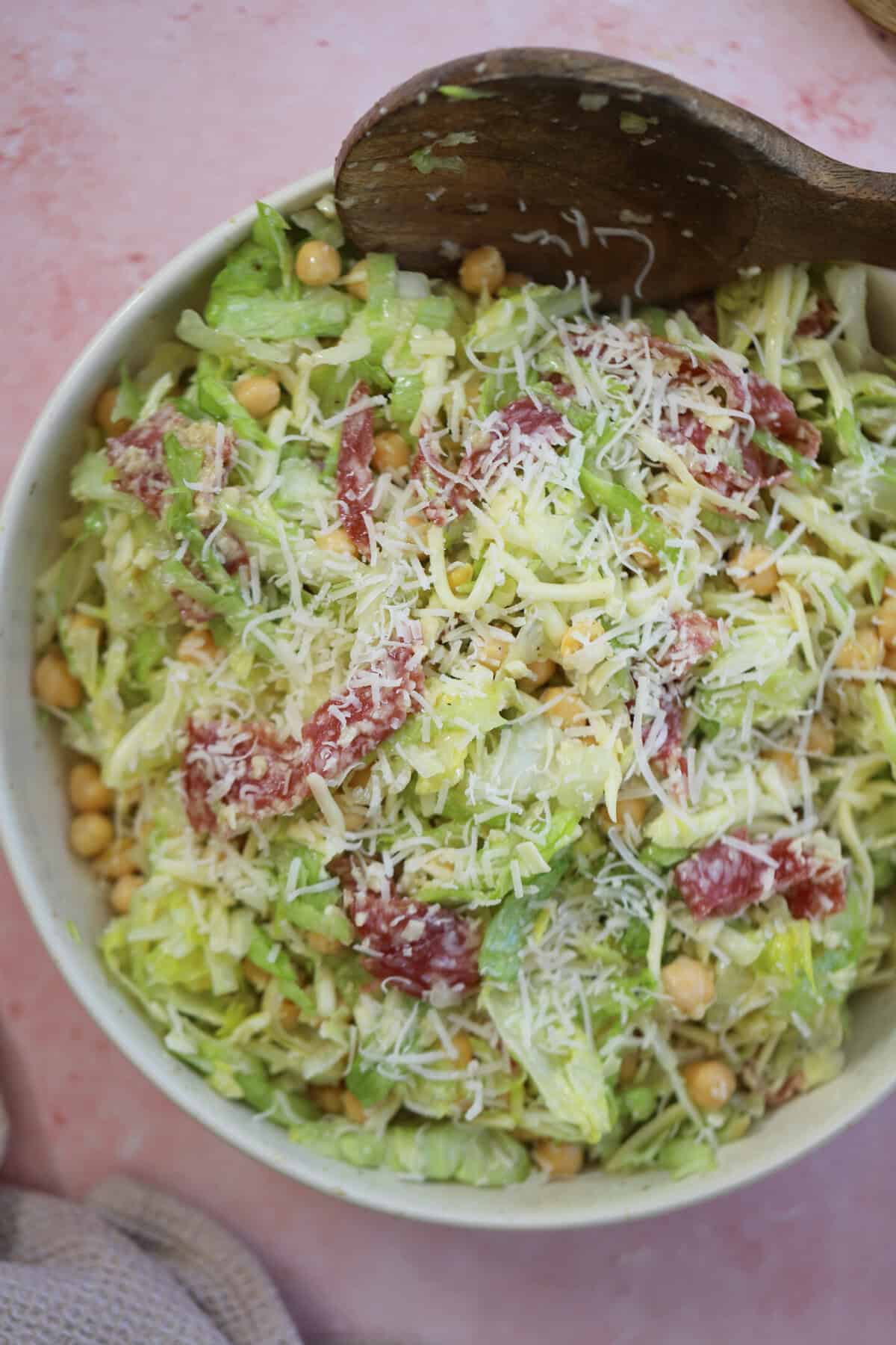 A bowl of La Scala salad with lettuce, shredded mozzarella, salami and chickpeas with a wooden serving spoon.