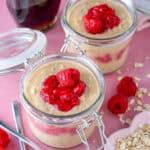 Two jars of PB&J Overnight Oats, topped with raspberries, with maple syrup behind.