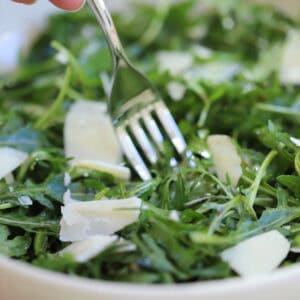 A hand holding a fork, taking a forkful from a bowl of rocket salad with parmesan shavings.