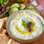 A bowl of tzatziki sauce with olives on top and cucumbers ready to dip.