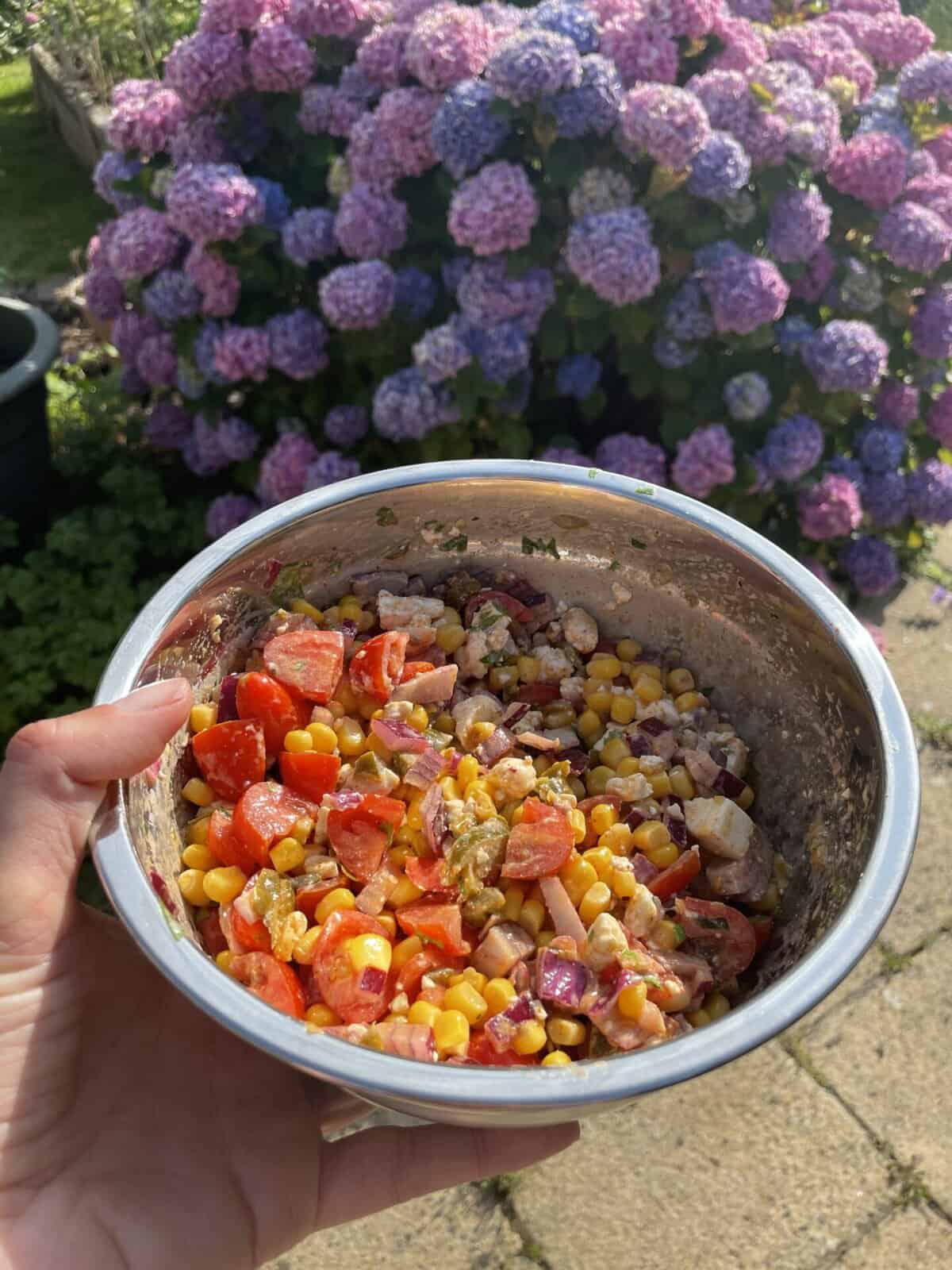 Hand holding a bowl of Mexican corn salad in front of a hydrangea bush.