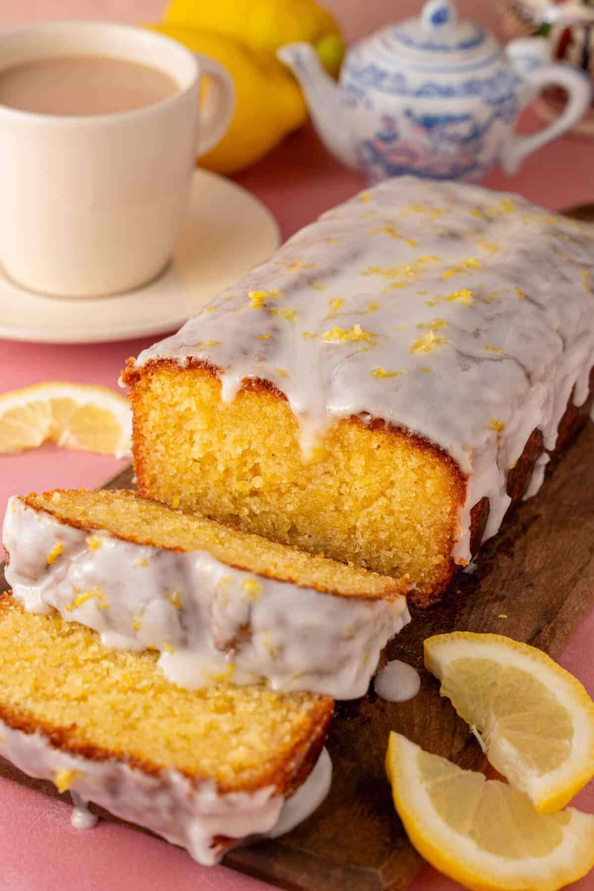 A gluten free lemon drizzle cake cut into slices with a cup of tea behind.