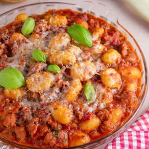 A sausage gnocchi bake in a glass bowl topped with cheese and basil leaves.