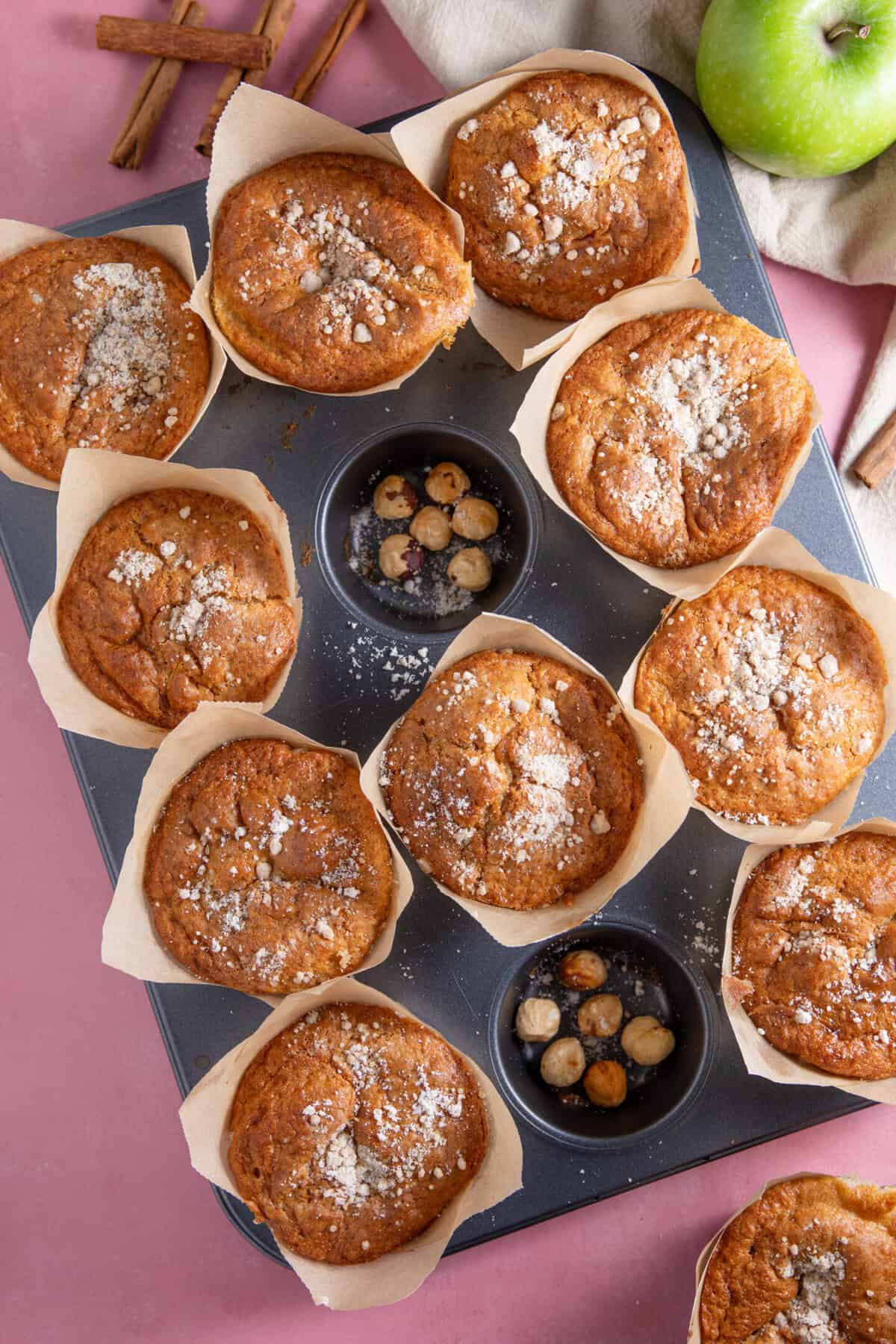 A tray of gluten free apple muffins surrounded by apples and hazelnuts.