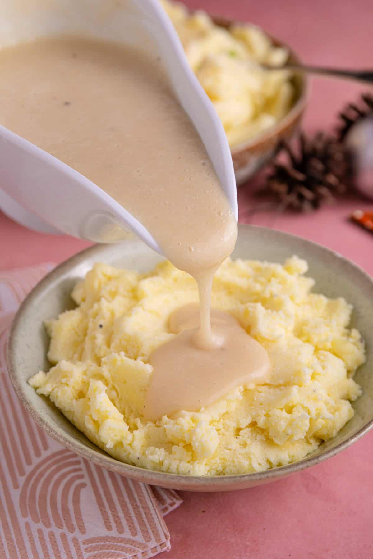 A jug of gravy pouring over a bowl of mashed potato.