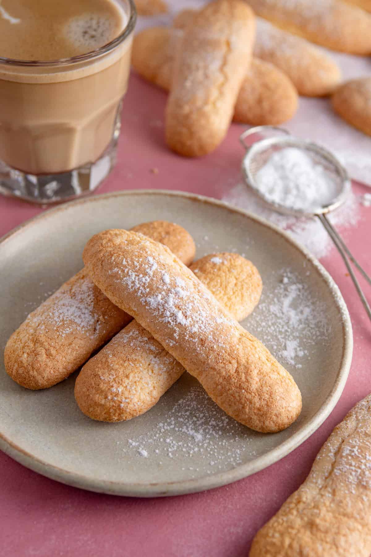 A plate of gluten free ladyfingers with a coffee behind.