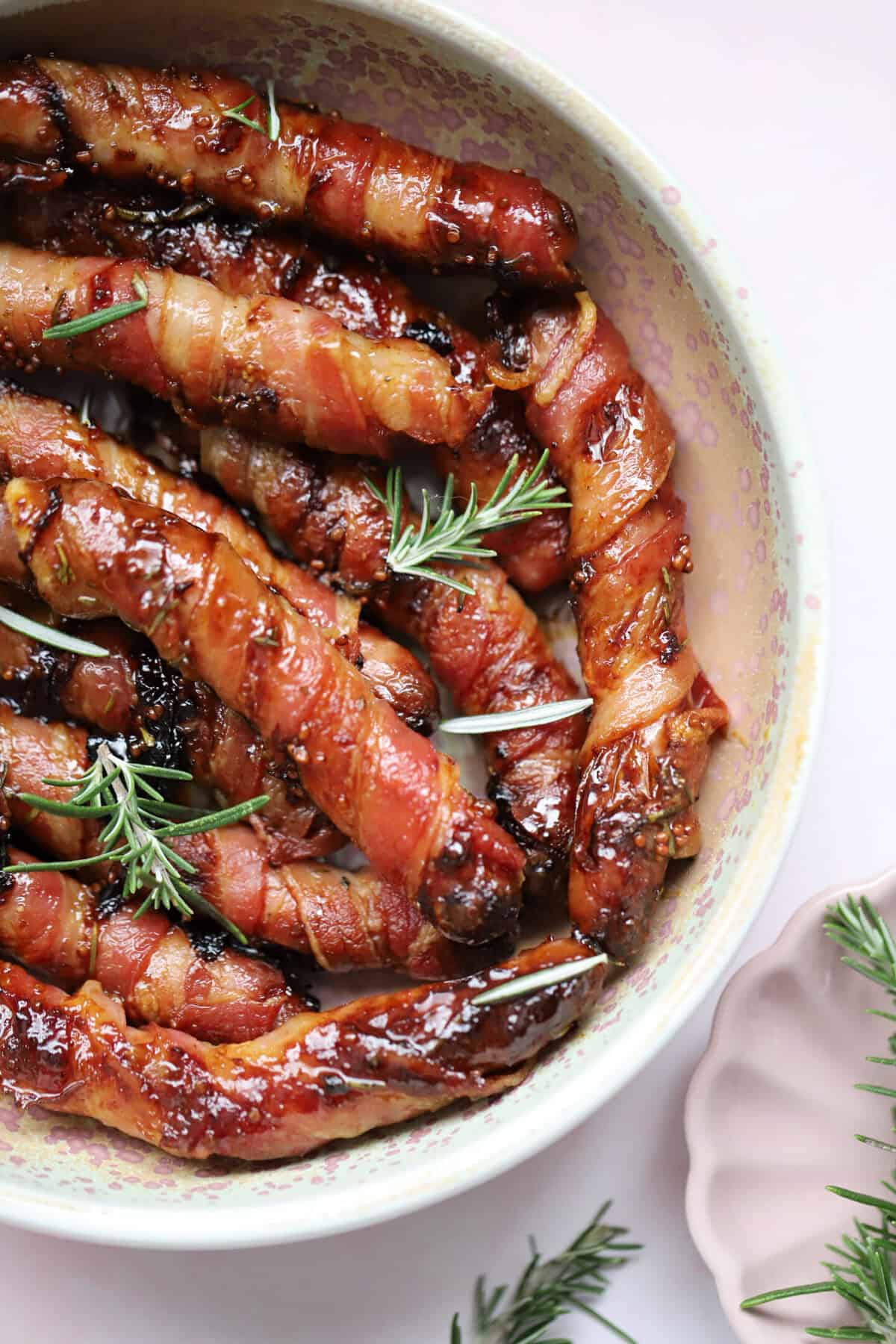 Close up of a bowl of gluten free pigs in blankets with rosemary sprigs.