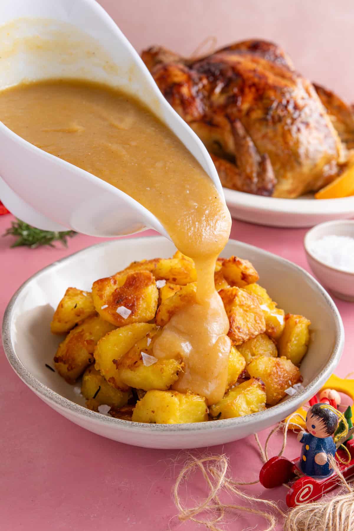 Gravy being poured over a plate of crispy gluten free roast potatoes with a roast chicken in the background.