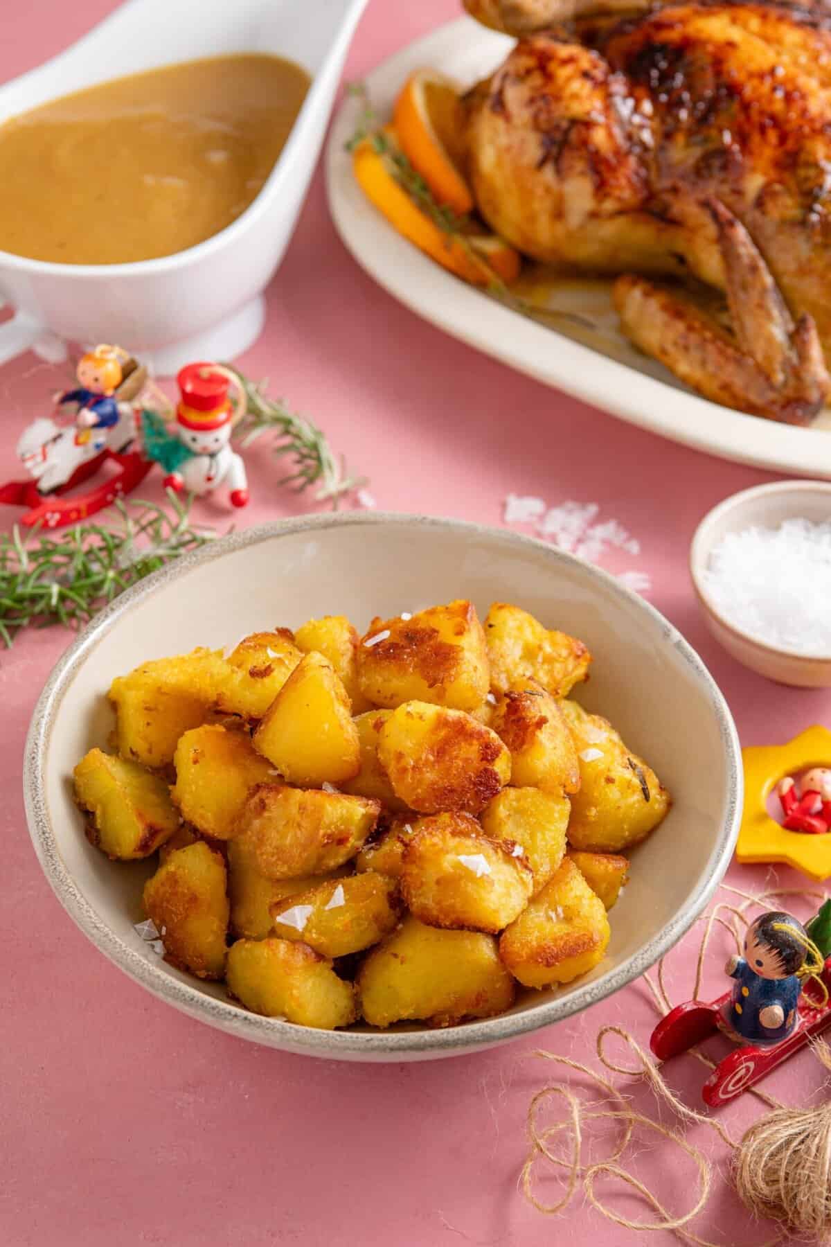 A bowl of crispy roast potatoes with gluten free gravy, a roast chicken and Christmas decorations.