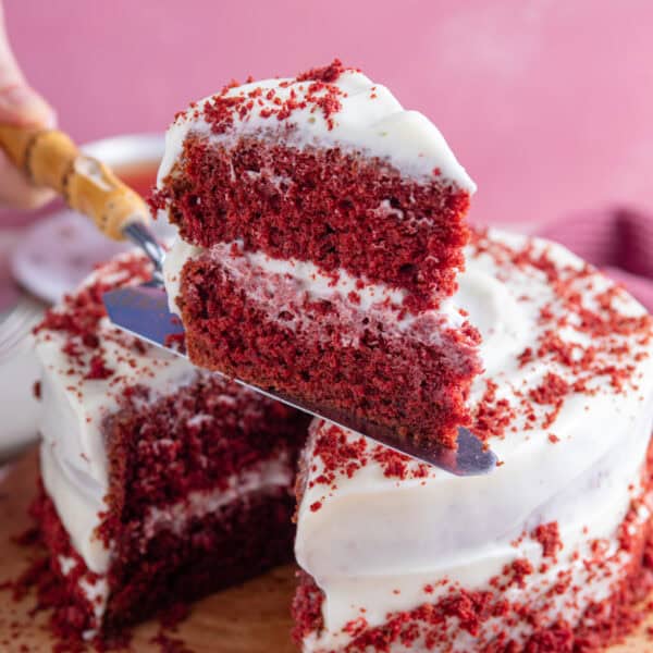 A slice of gluten free red velvet cake on a cake slice with the cake behind it.