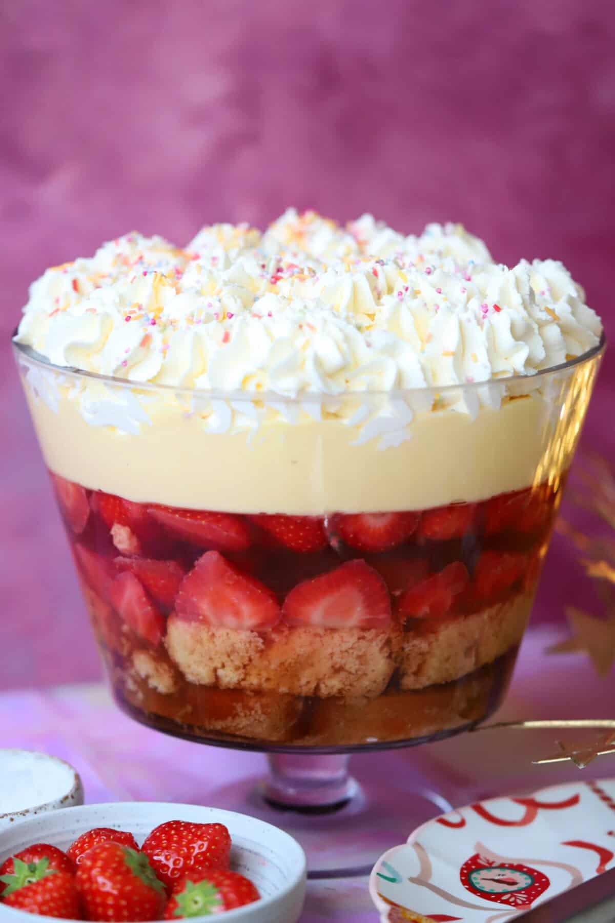A gluten free trifle in a glass trifle bowl with a serving spoon in front and lights behind.