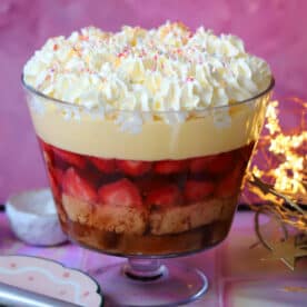 A gluten free trifle in a glass trifle bowl with a serving spoon in front and lights behind.