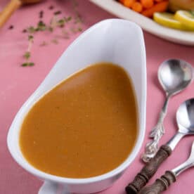A jug of gluten free gravy with a plated up roast dinner in the background.