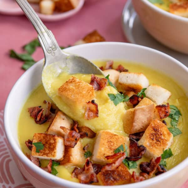 A bowl of parsnip soup topped with crispy bacon and croutons, with a spoon.