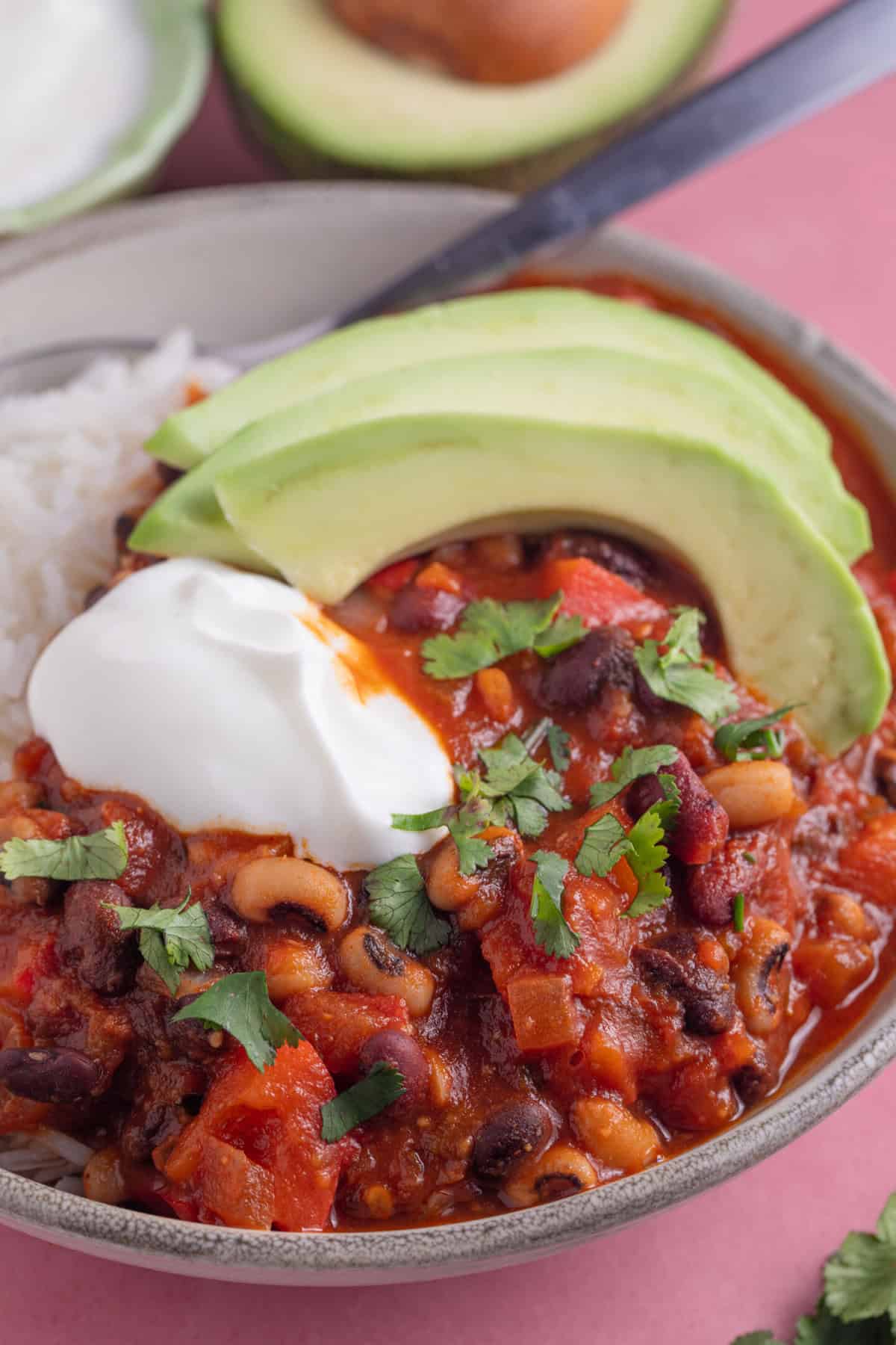 A bowl of three bean chilli with rice, sliced avocado and soured cream.