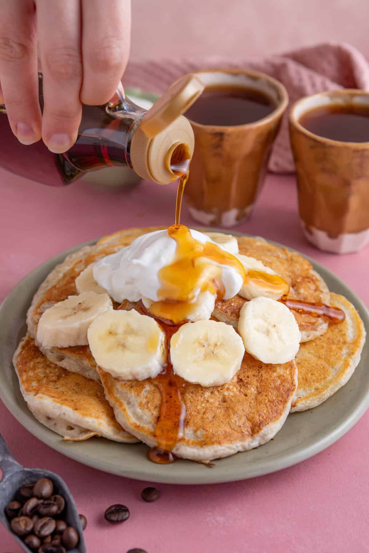 Hand pouring a bottle of syrup over a plate of gluten free banana pancakes with Greek yoghurt and sliced banana.