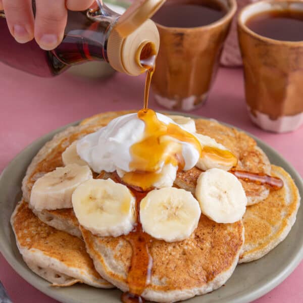 Hand pouring a bottle of syrup over a plate of gluten free banana pancakes with Greek yoghurt and sliced banana.