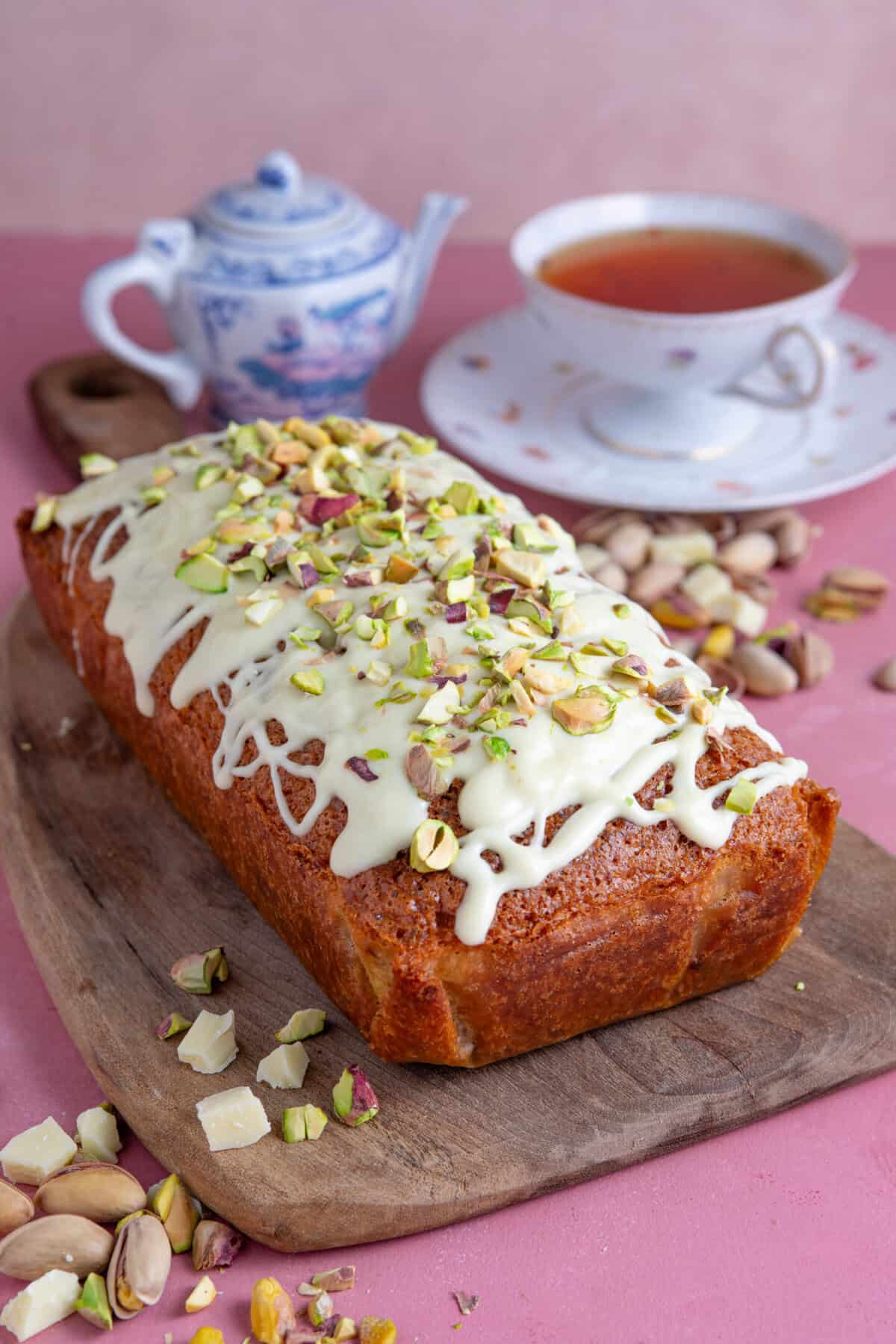 A gluten free pistachio loaf cake on a board with a tea set behind.