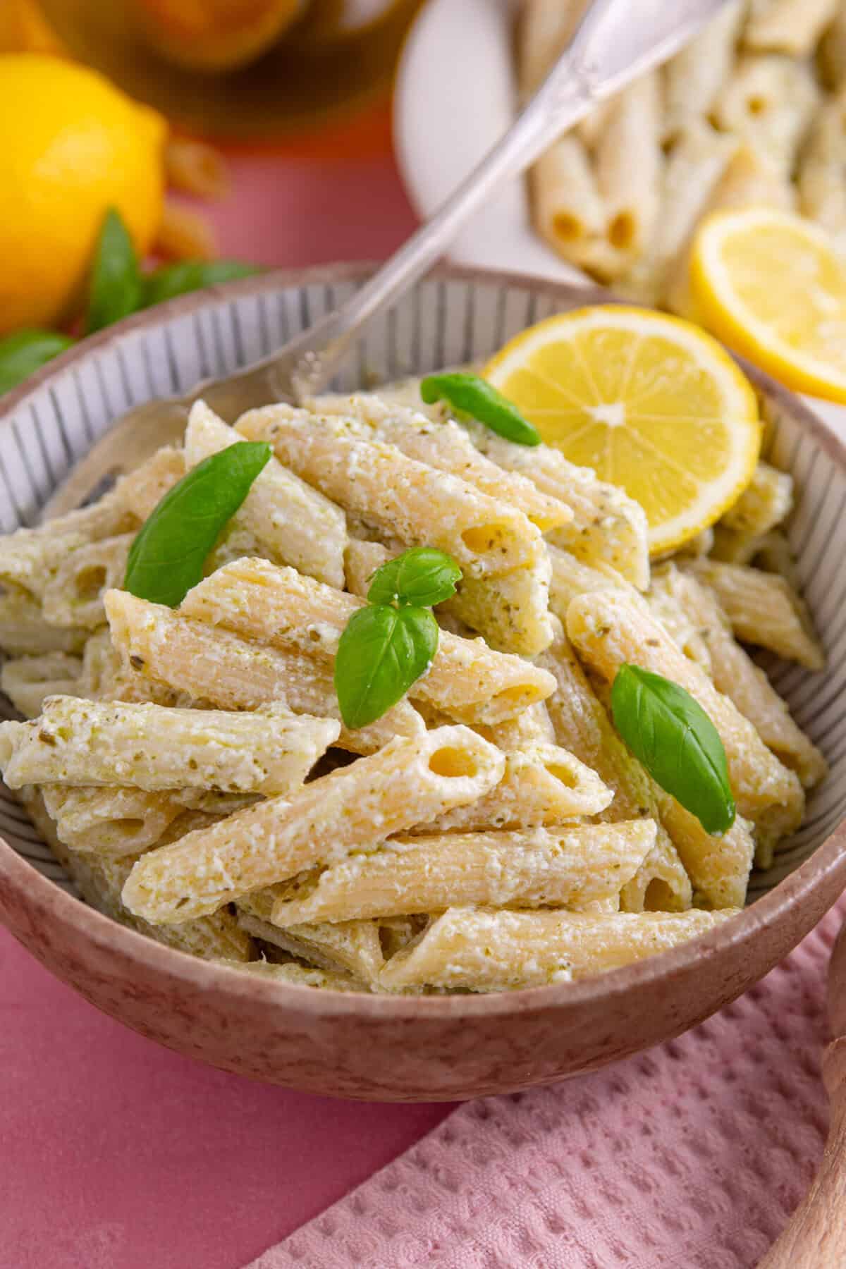 A bowl of creamy ricotta and pesto pasta with basil and a lemon slice.