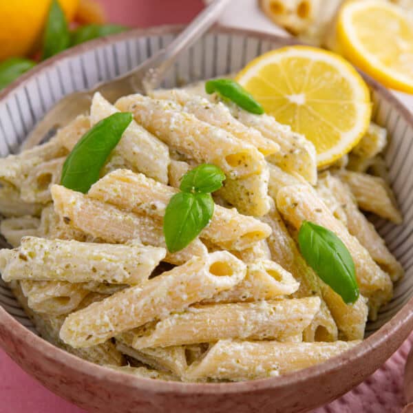 A bowl of creamy ricotta and pesto pasta with basil and a lemon slice.