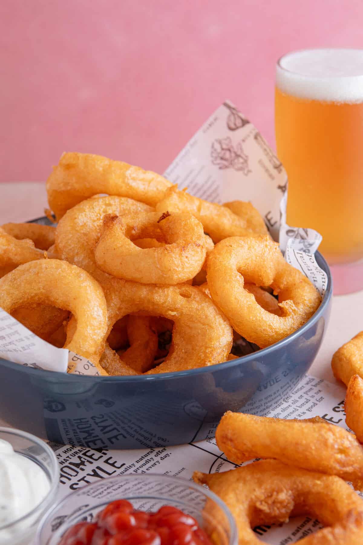 A bowl of gluten free onion rings with a pint of gluten free lager in the background.