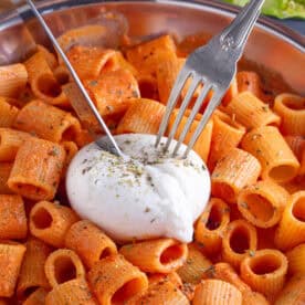A knife and fork cutting into a ball of burrata on top of a creamy tomato pasta dish.