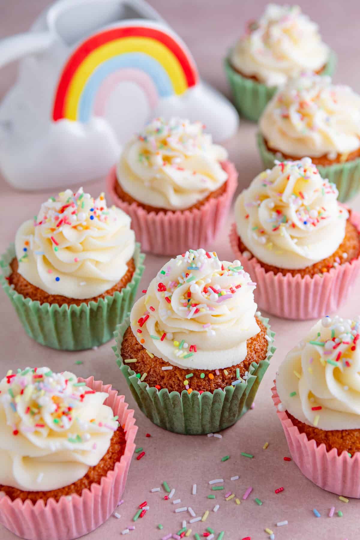 Gluten free vanilla cupcakes on a pink worktop with frosting and sprinkles on top.
