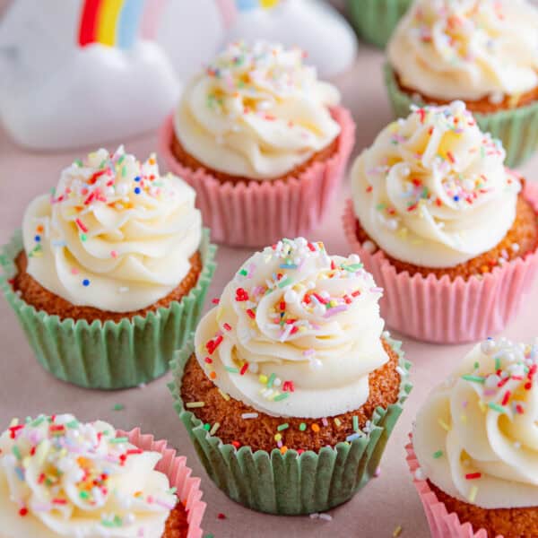 Gluten free vanilla cupcakes on a pink worktop with frosting and sprinkles on top.