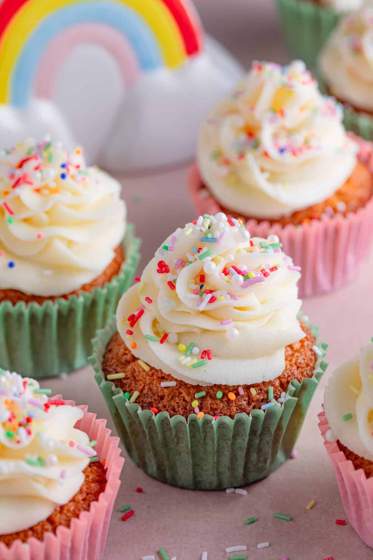 Gluten free vanilla cupcakes on a pink worktop with frosting and sprinkles on top.