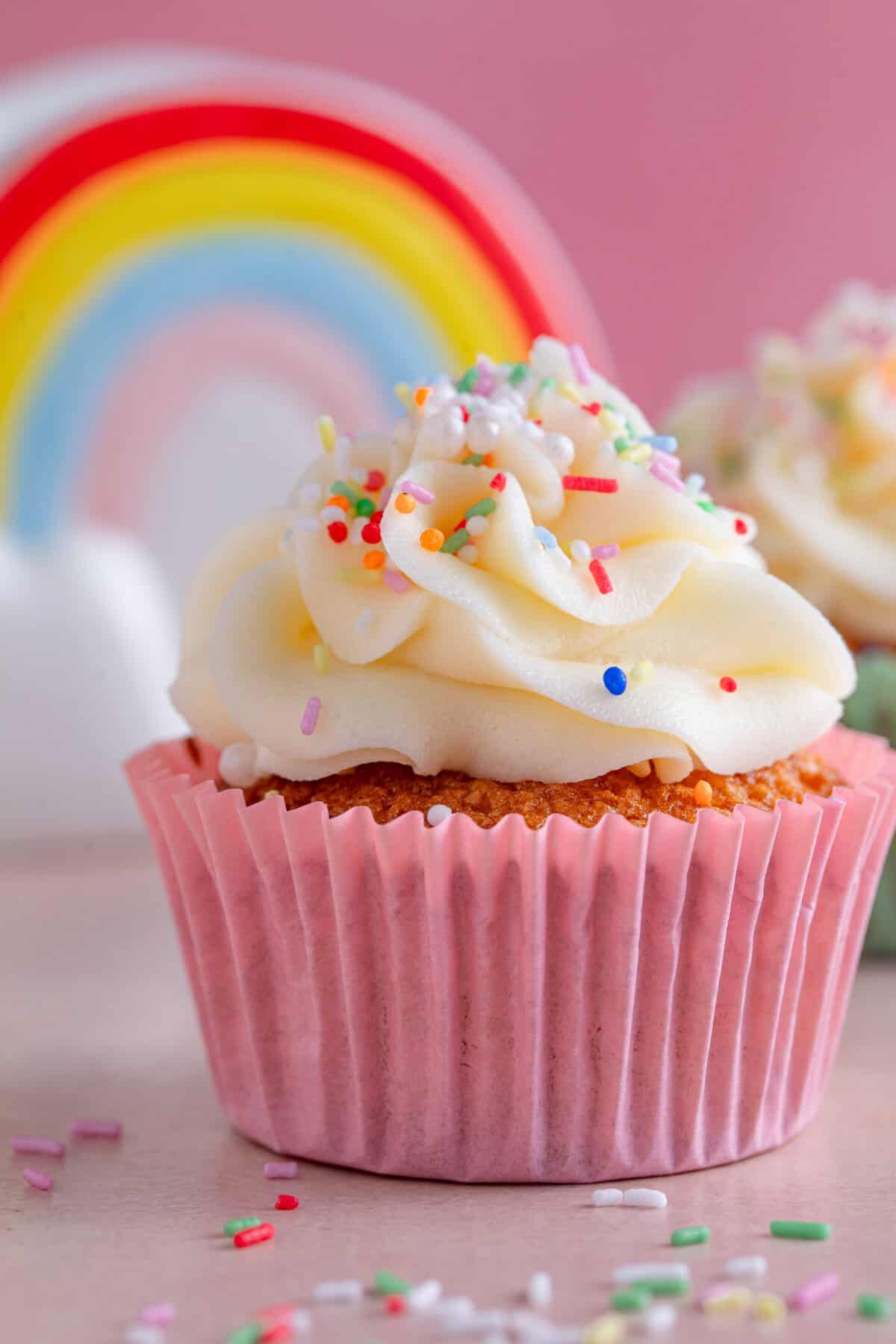Close up of a vanilla cupcake in a pink case with a rainbow mug behind.