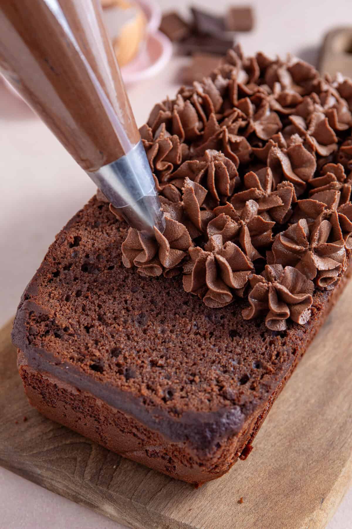 Process shot of a gluten free chocolate loaf cake recipe showing the frosting being pipped onto the cake in swirls.