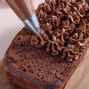 Process shot of a gluten free chocolate loaf cake recipe showing the frosting being pipped onto the cake in swirls.