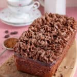 A gluten free chocolate loaf cake with chocolate frosting and a tea set in the background.