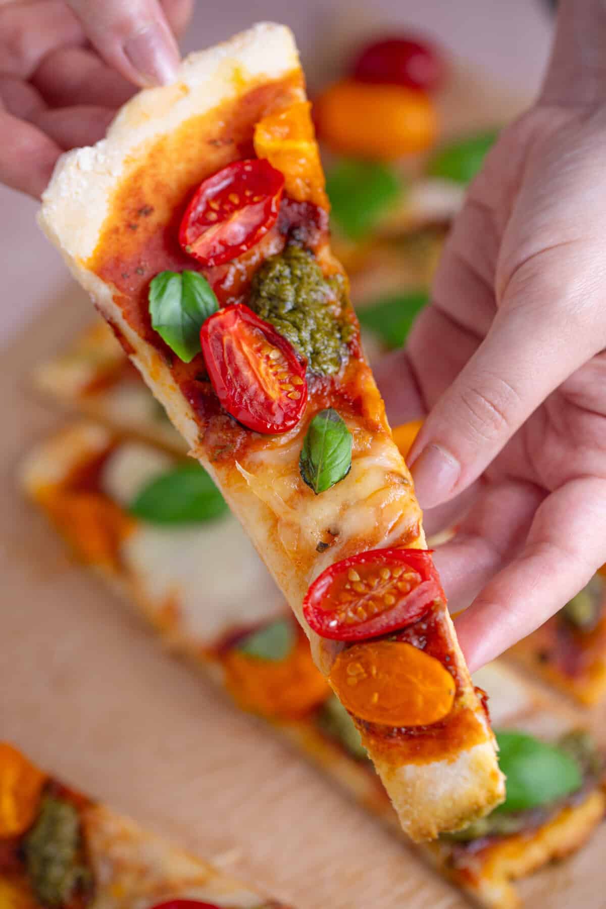 Hands holding a strip of gluten free flatbread pizza with tomatoes and pesto.
