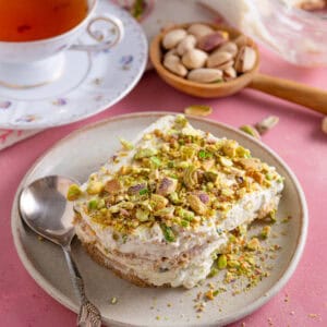 A slice of pistachio tiramisu on a plate with a cup of tea and pink worktop.