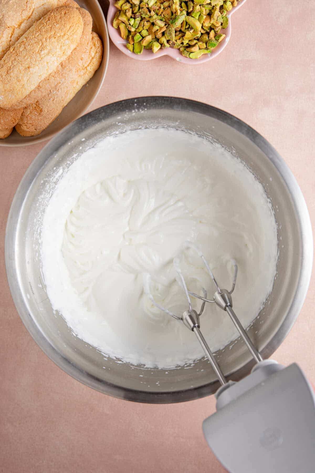 Process shot of a pistachio tiramisu recipe showing the double cream being whisked in a large bowl.