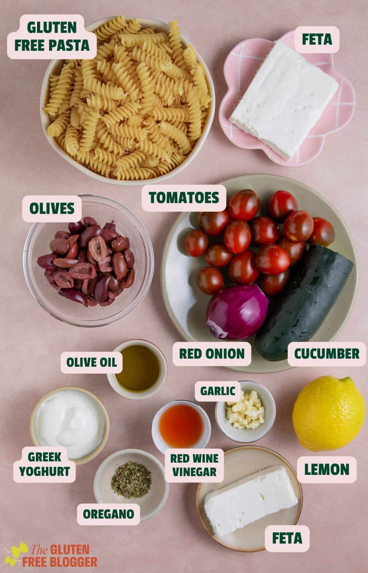 Ingredients for a Greek pasta salad with text labels, laid out on a pink work surface.