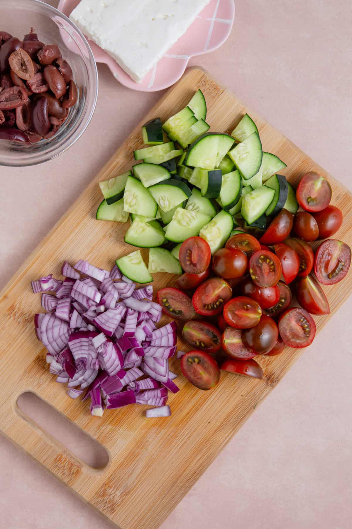 Process shot of a Greek pasta salad recipe showing tomatoes, cucumber and red onion diced up on a chopping board.