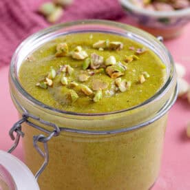 A jar of homemade pistachio cream with a pink backdrop, with crushed pistachio sprinkled on top.