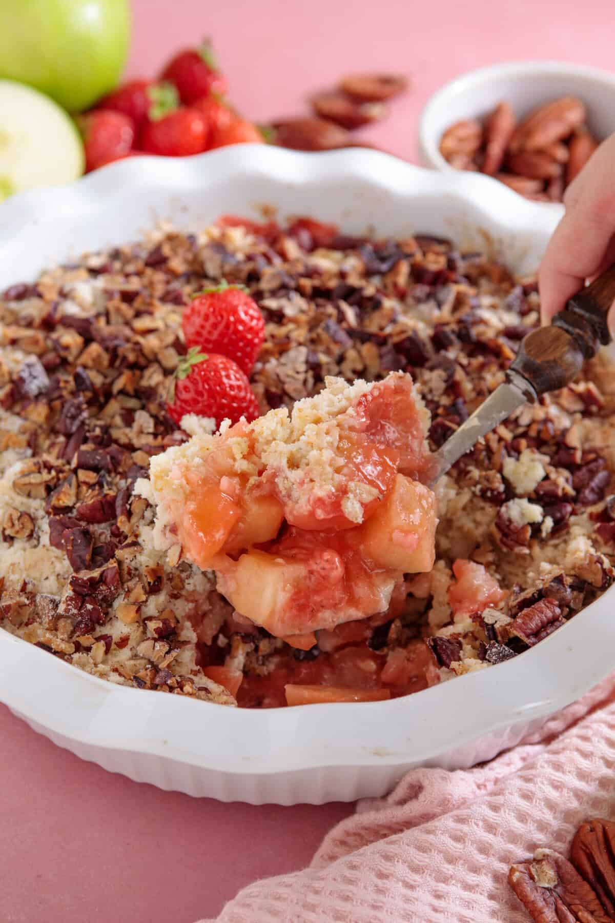An apple and strawberry crumble with a spoonful being lifted out of the tin.