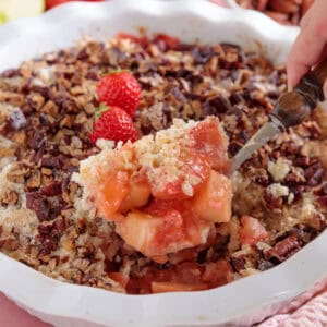An apple and strawberry crumble with a spoonful being lifted out of the tin.