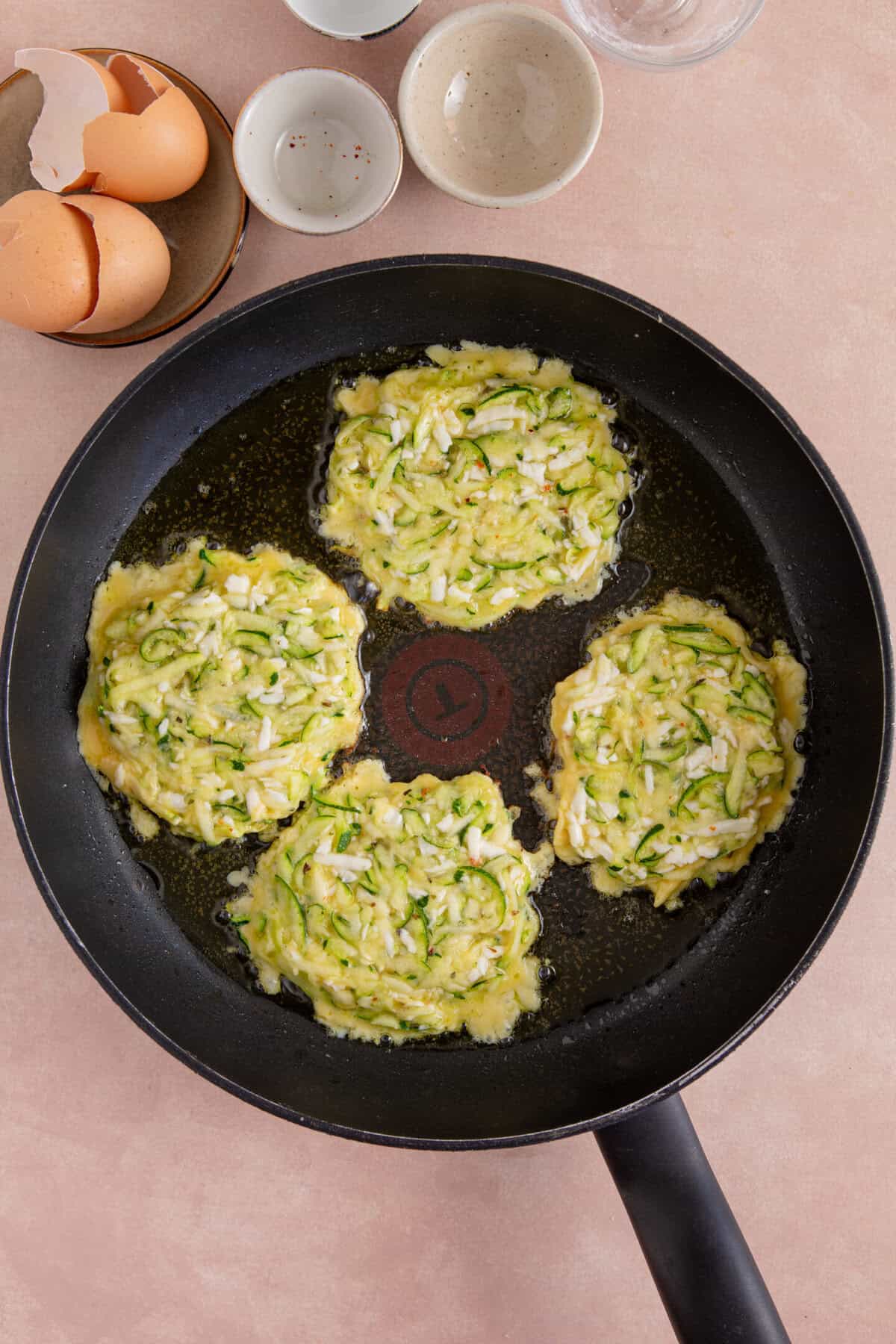 Process shot of courgette and halloumi fritters showing four fritters in a pan, cooking on one side before flipping.