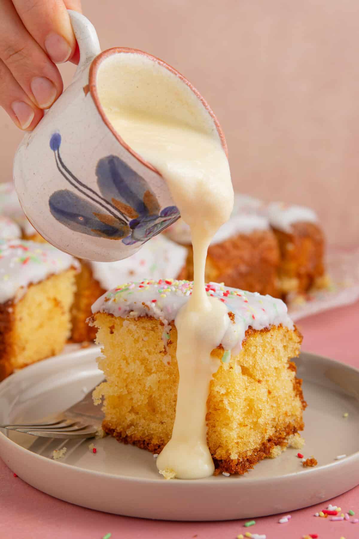 A just of custard being poured over a square of school dinner cake with white icing and rainbow sprinkles.