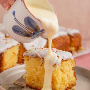 A jug of custard being poured over a square of school dinner cake with white icing and rainbow sprinkles.